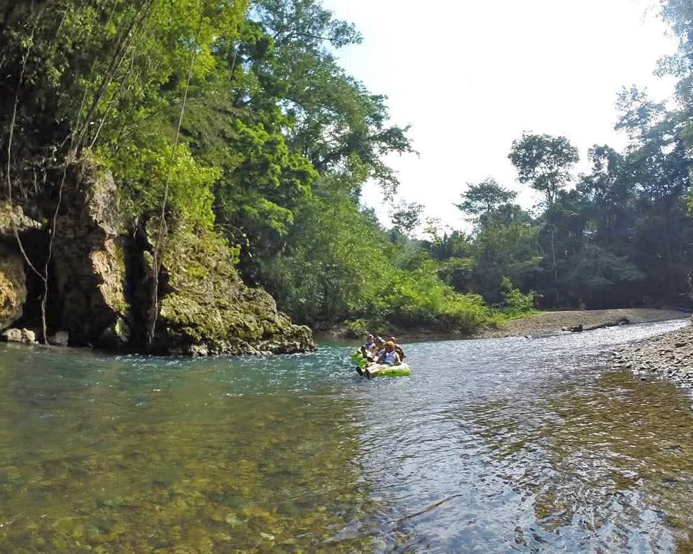 Two people in an inflatable raft float down a tranquil river surrounded by lush greenery.