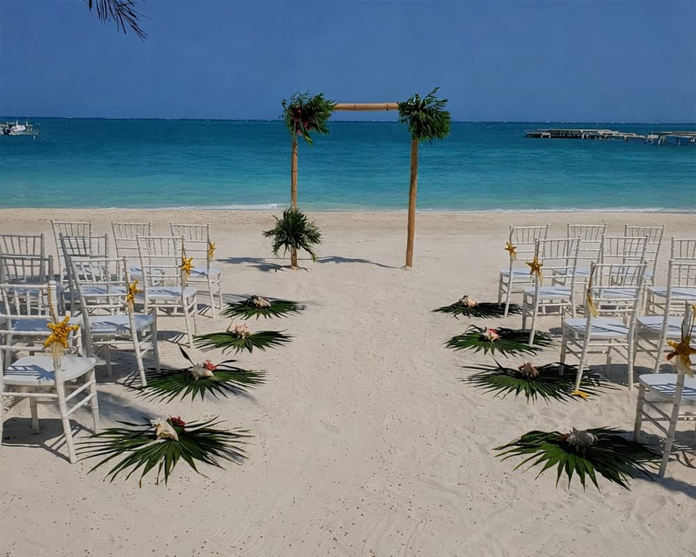 Beach wedding setup with chairs arranged on sand facing a floral archway and the ocean.