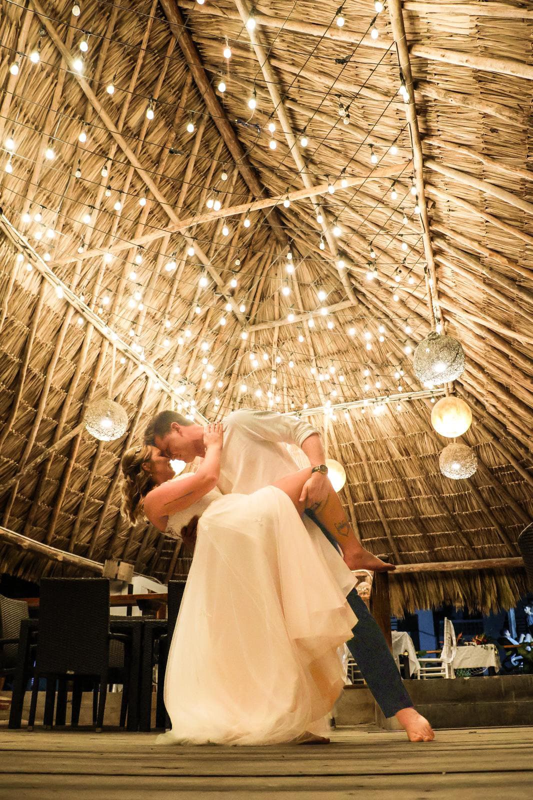 A couple dances joyfully under a beautifully lit thatched roof.