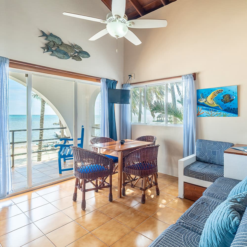 A bright coastal living area with a dining table, blue chairs, and large windows overlooking the ocean.