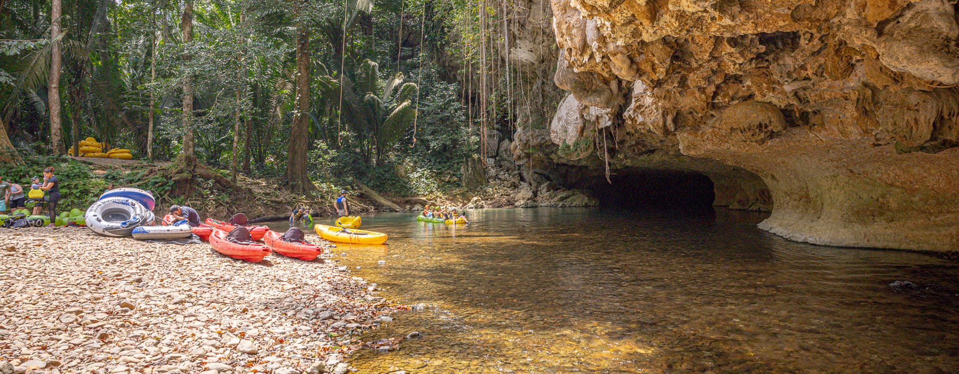 Kayakers and tubers relax near a rocky shore and cave entrance in a lush, tropical setting.
