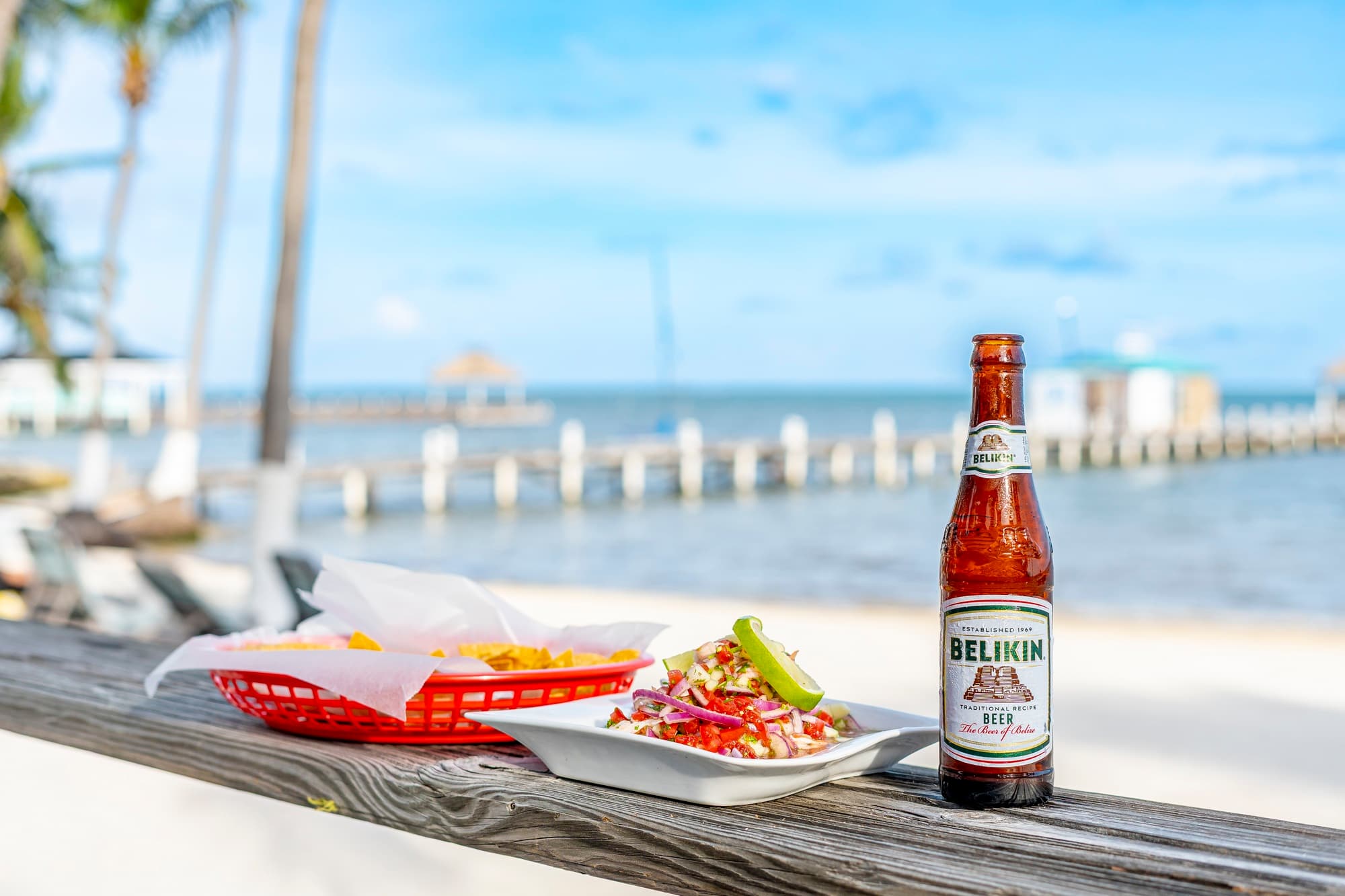 A bottle of Belikin beer next to a plate of seafood and a basket of fries, with a scenic waterfront in the background.