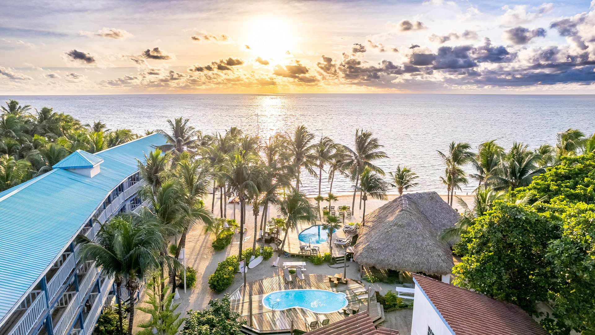 Aerial view of a coastal resort with palm trees, pools, and a sunset over the ocean.