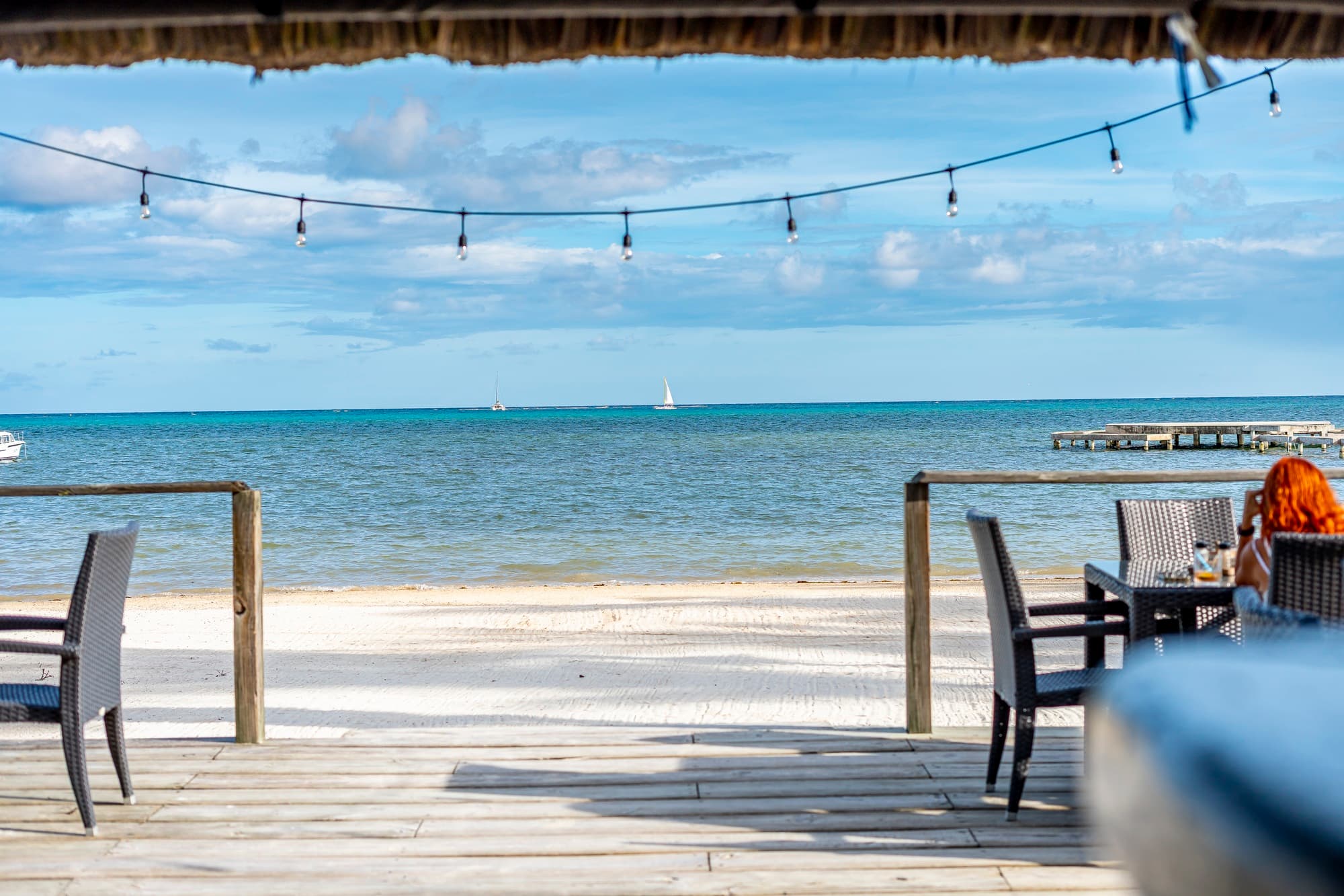 A scenic view of a beach with wooden decks, chairs, and a distant sailboat under a blue sky.