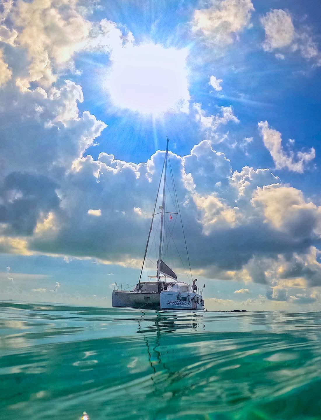 A sailboat floats on calm water beneath a bright sun and scattered clouds.