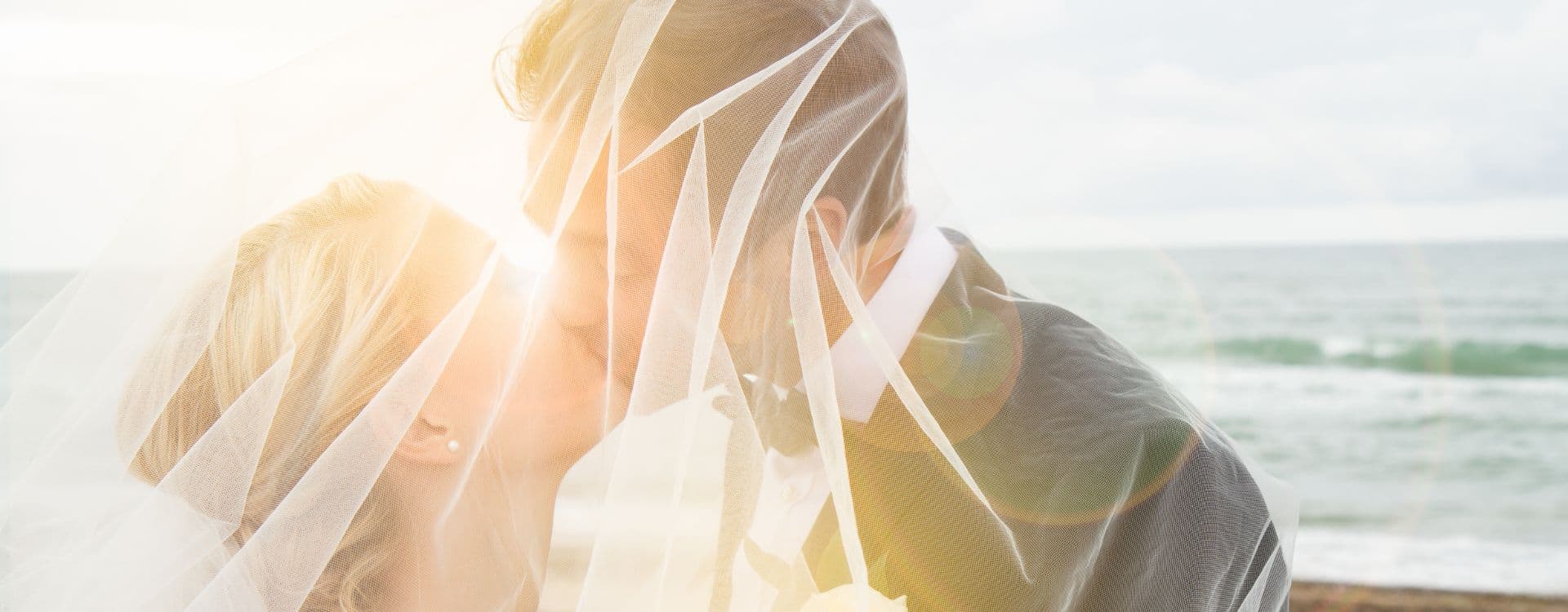 A couple kisses under a veil at the beach with sunlight shining through.