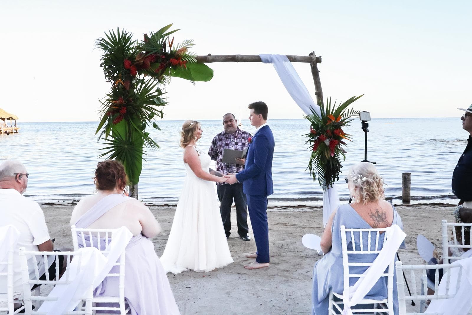 A couple stands at a floral arch on the beach, exchanging vows during their wedding ceremony.