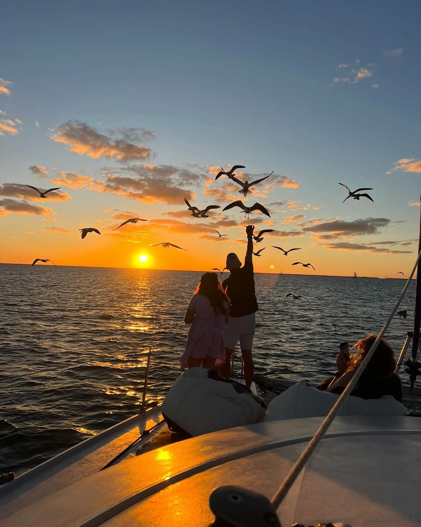 A silhouette of two people on a boat watching birds fly over a sunset on the water.